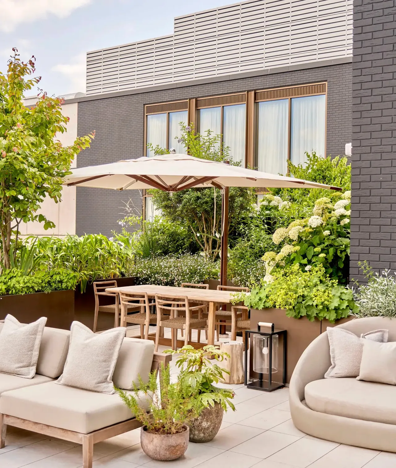 A planted garden terrace with sofas, a parasol and a dining table.