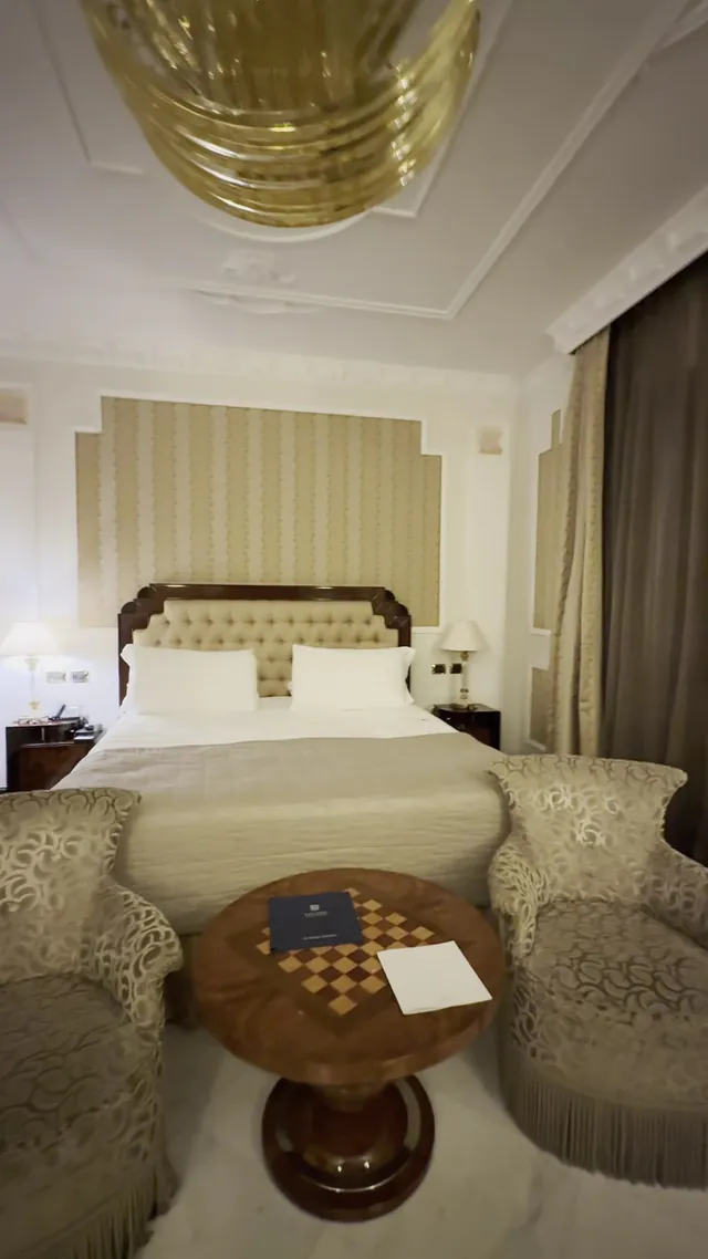 A classic guest room with a padded headboard, armchairs and a small round table.