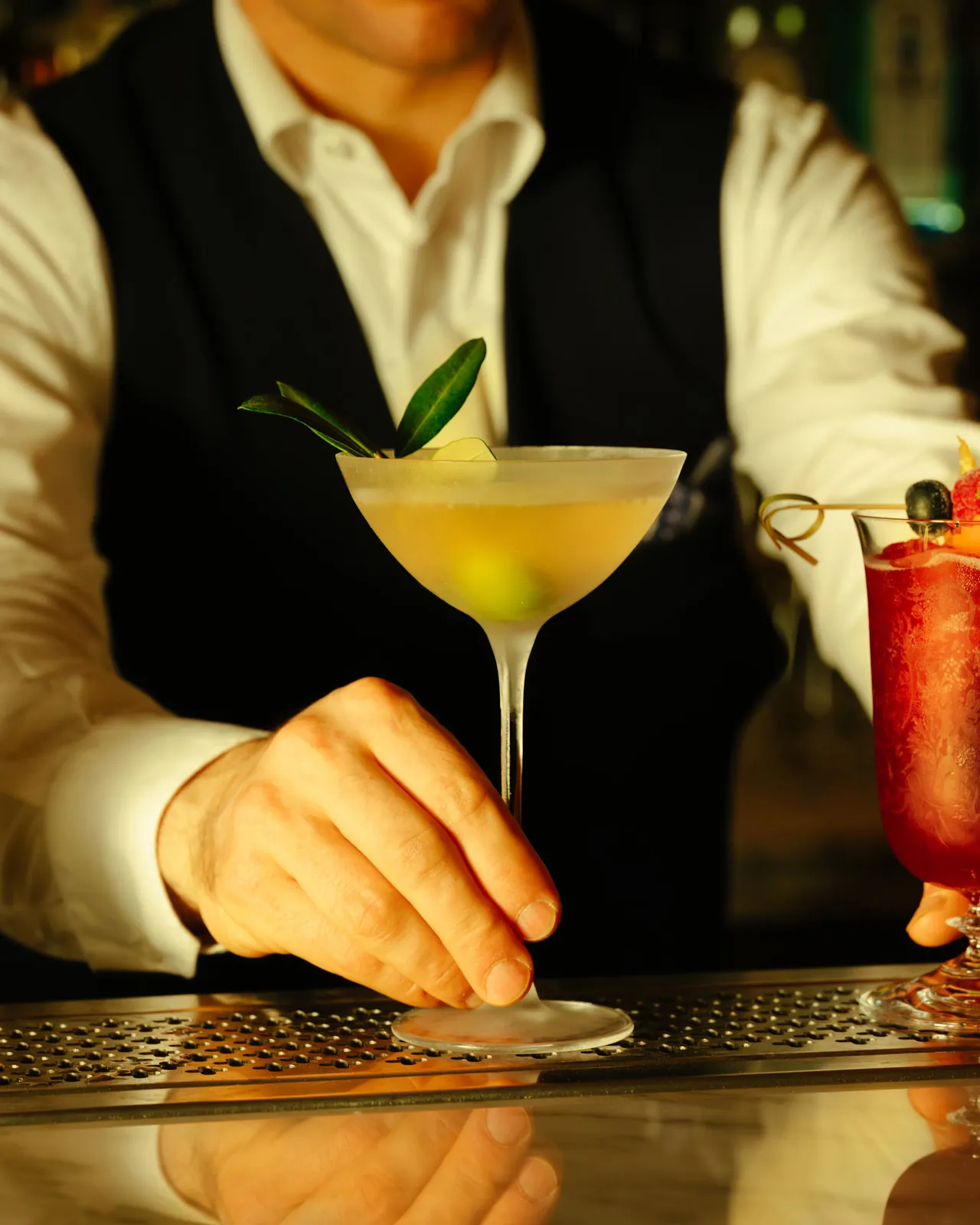 A bartender sets down a pale lemon cocktail garnished with an olive sprig.