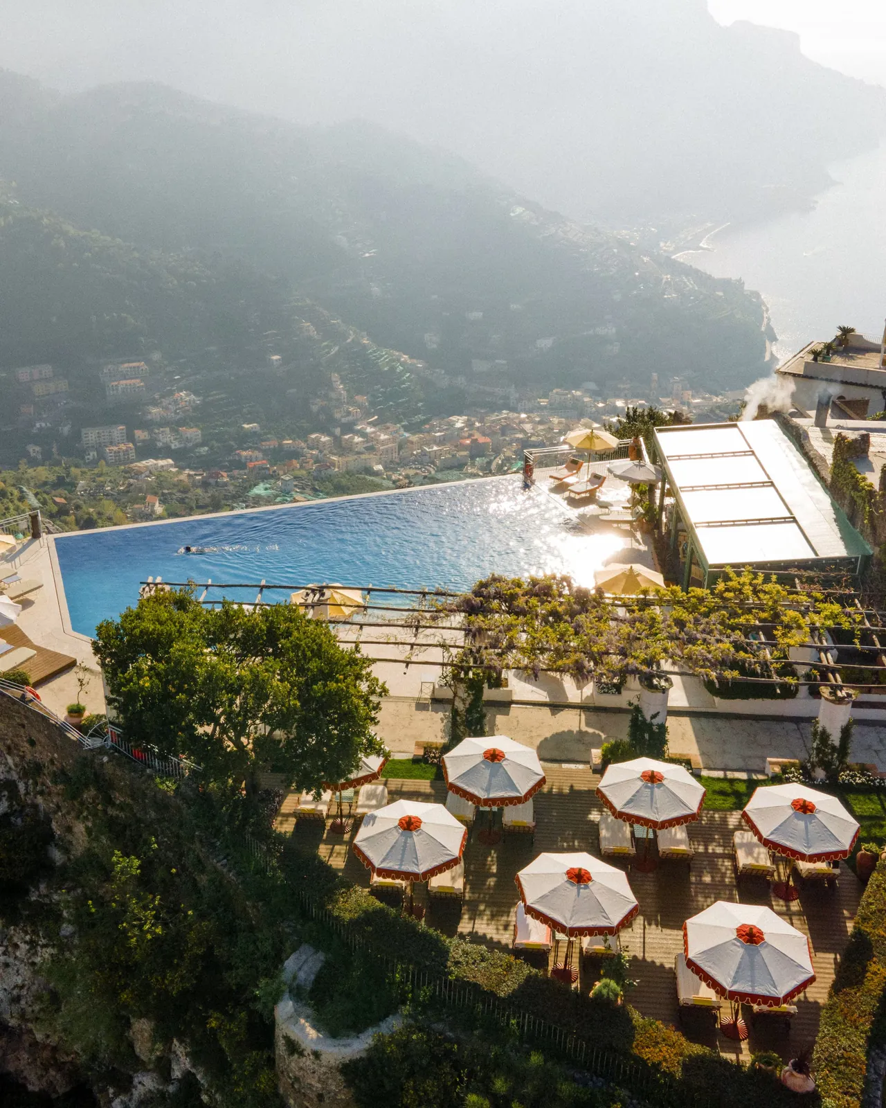 The infinity pool and a terrace of white parasols seen from above, with the coast below.