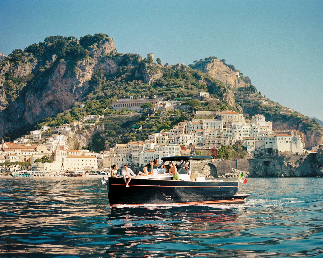 A black wooden boat cruises below a cliffside town on the coast.