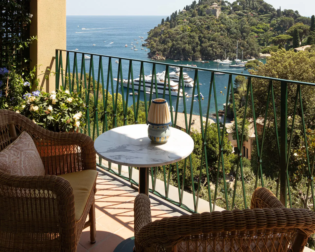 A table and wicker chairs on a room terrace above the bay and its moored boats.