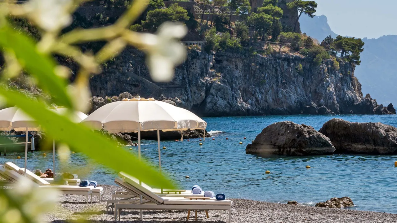 Parasols and loungers on the pebble beach, with rocks in the clear water.