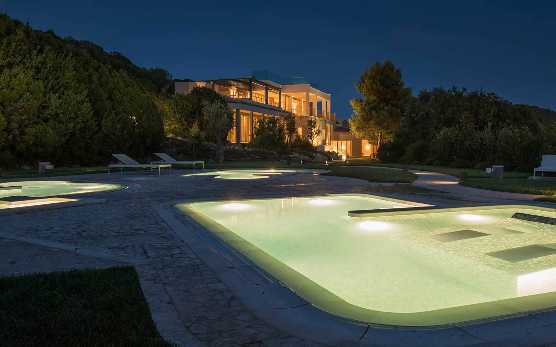Lit pools at Chia Laguna Resort, Sardinia, at night with a glowing villa and dark trees behind