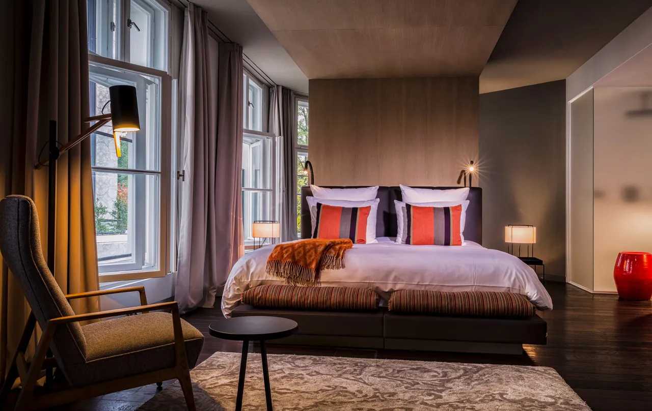 A suite bedroom with tall windows, a reading lamp and orange accent cushions.