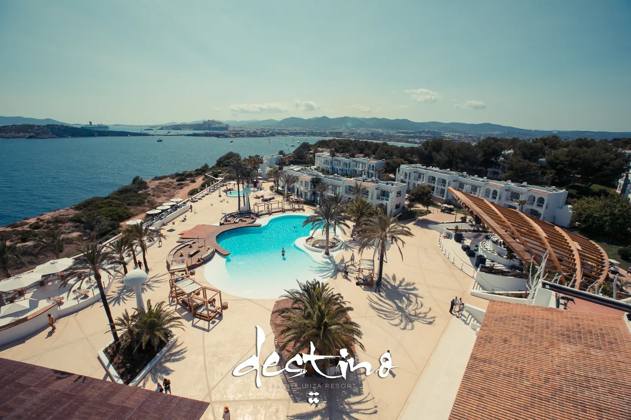 Aerial view of the pool, palms and white buildings of Destino Five Ibiza above the bay