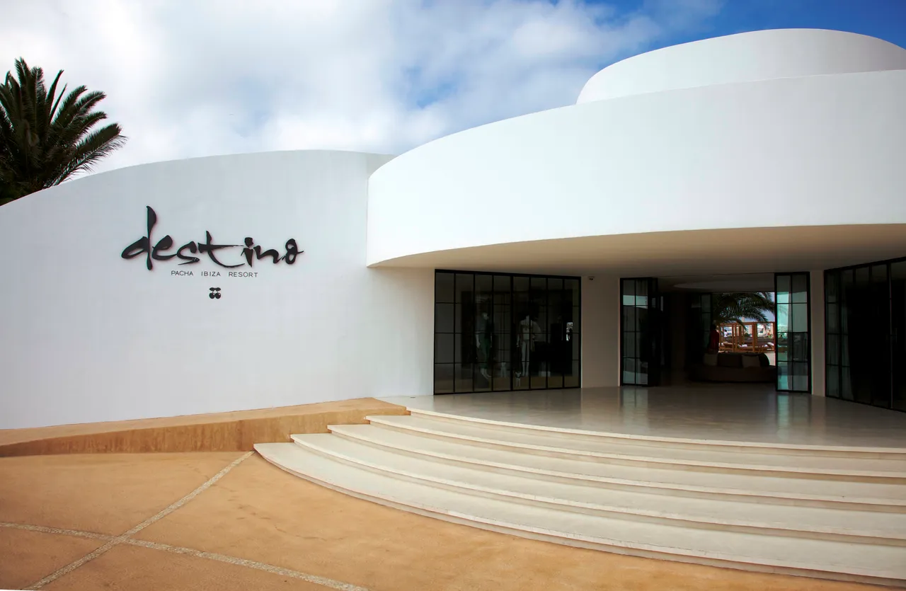 Curved white entrance building of Destino Five Ibiza with wide stone steps, glass doors and a palm tree