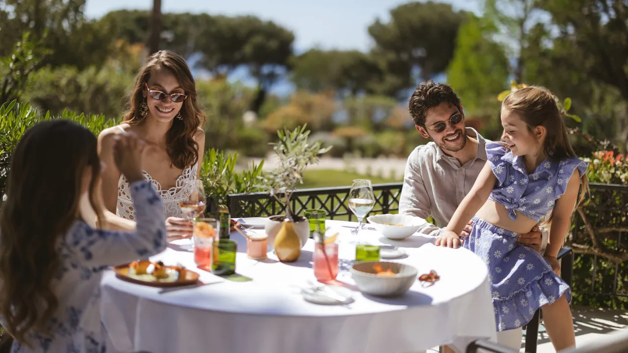 A family at lunch at a white-clothed table on a sunny garden terrace.