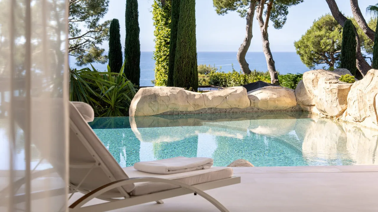 A sun lounger beside a private pool, with pines and the sea beyond the rocks.