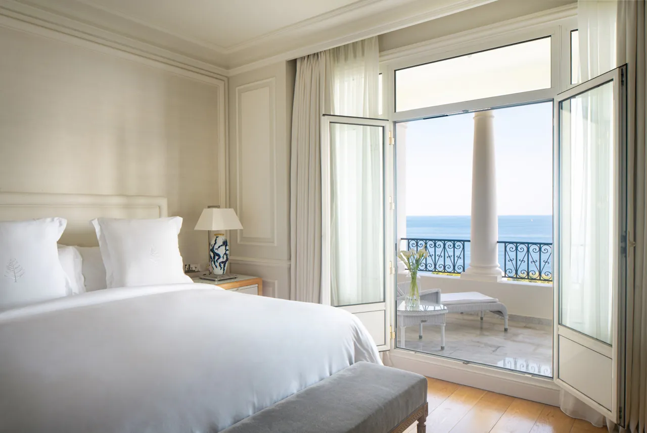 A light bedroom with tall French windows open onto a sea-view balcony.