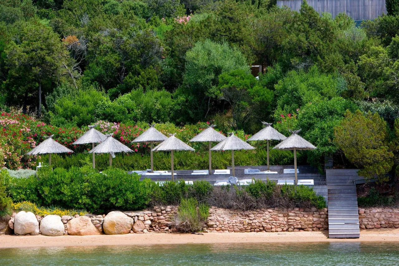 White parasols and loungers line the private beach and wooden jetty below the green hillside.