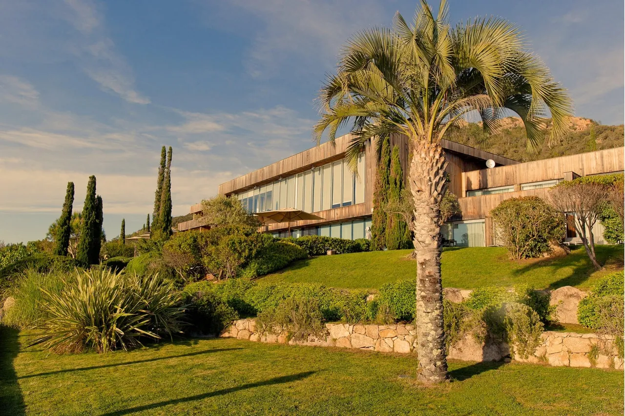 The low wood and glass hotel building behind palms and cypress trees in the garden.