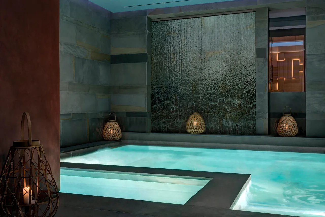 The softly lit spa pool in front of a dark stone water wall.