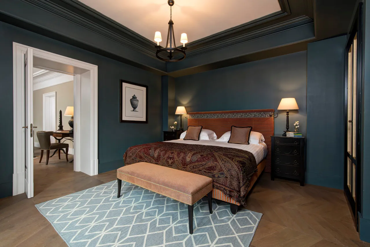 A dark teal bedroom with a chandelier and a velvet bench at the bed.