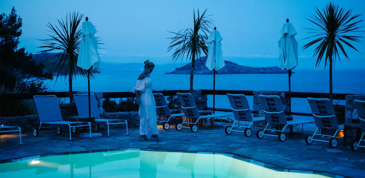 The pool terrace lit up at dusk with palm trees and the sea behind.
