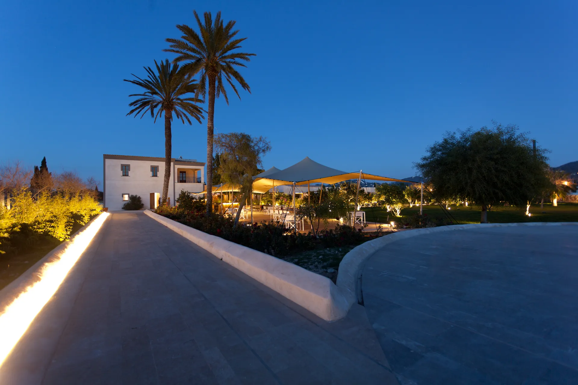 Lit pathway to Hotel Xereca, Ibiza, at dusk with tall palms, a white villa and a canopied terrace