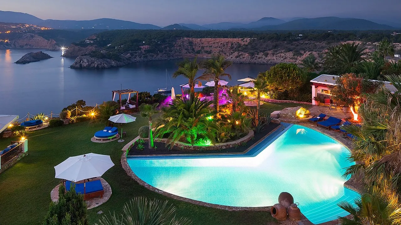 Pool at Las Brisas lit up at dusk, surrounded by lawns, palms and parasols above a calm bay