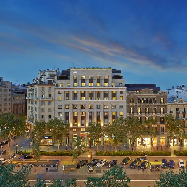 Lit stone facade of Mandarin Oriental, Barcelona, at dusk on a tree-lined boulevard with taxis