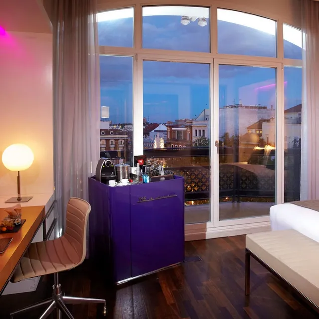 Room at ME Madrid with a purple minibar cabinet, desk lamp and tall balcony windows at dusk