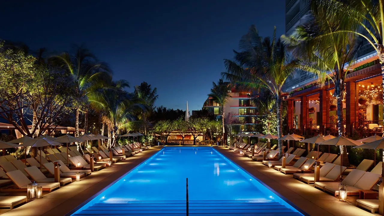 Long pool at The Miami Beach Edition lit blue at night, lined with loungers, parasols and palm trees