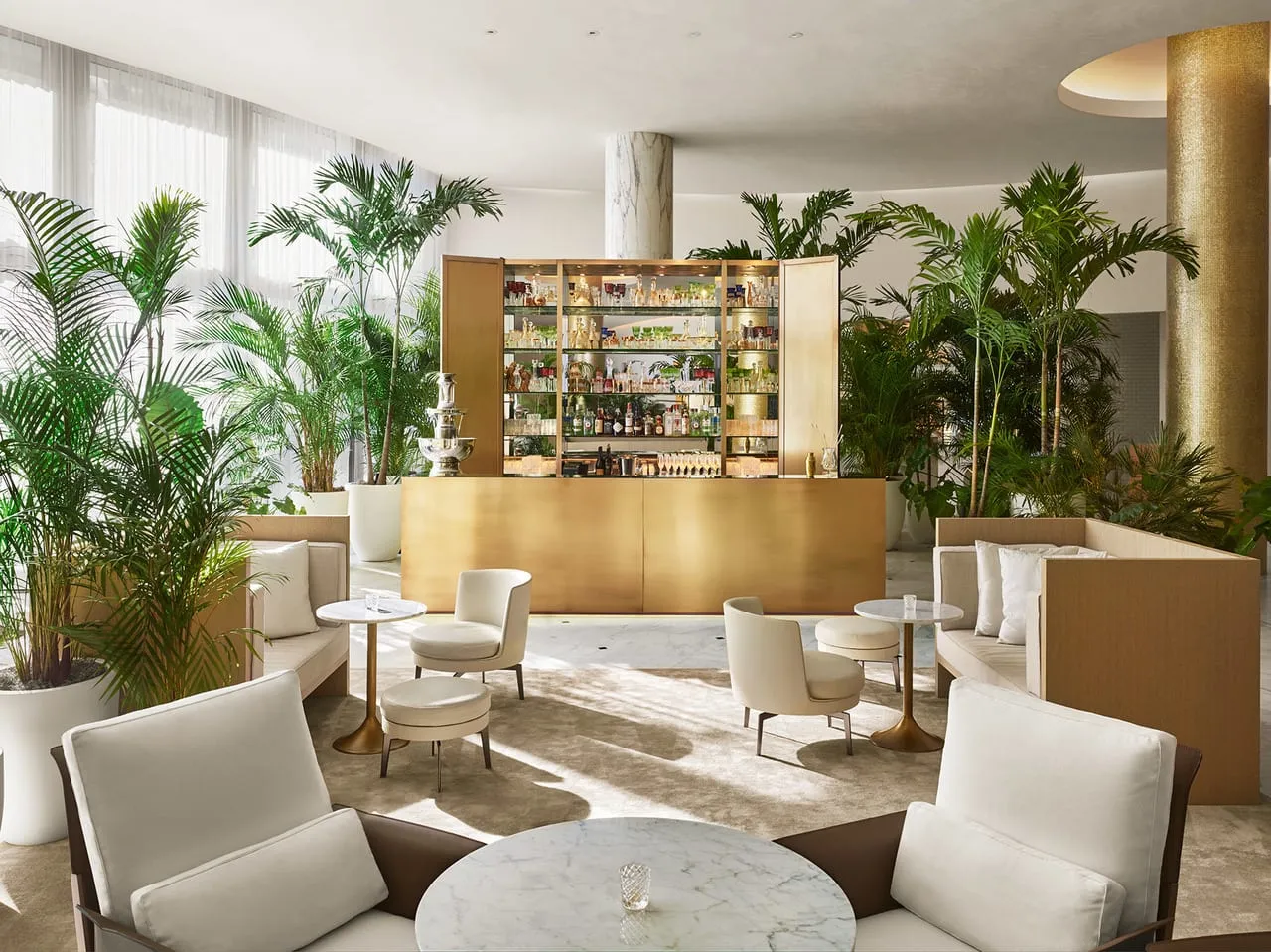Lobby bar at The Miami Beach Edition with a brass bar cabinet, white armchairs, marble tables and palms