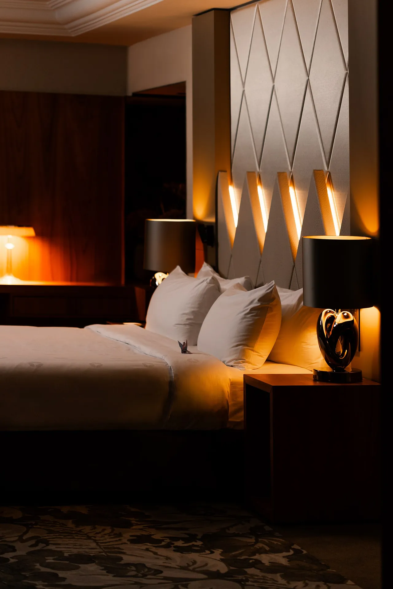 A bedroom in the evening, lit by bedside lamps and a glowing headboard.