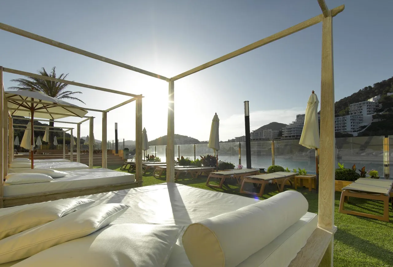 Canopy daybeds on a lawn terrace at Palladium Hotel Don Carlos with a sunlit bay and hills behind