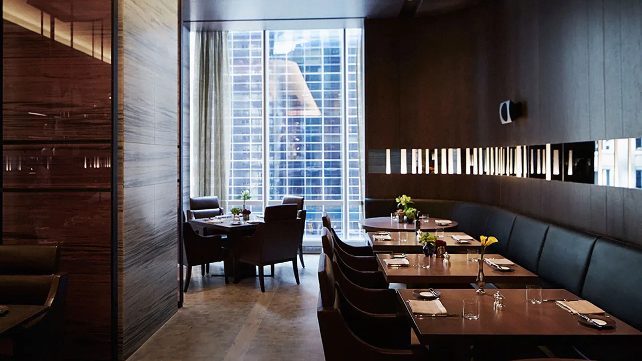 Restaurant at Park Hyatt New York with dark wood walls, banquettes and a tall window onto skyscrapers