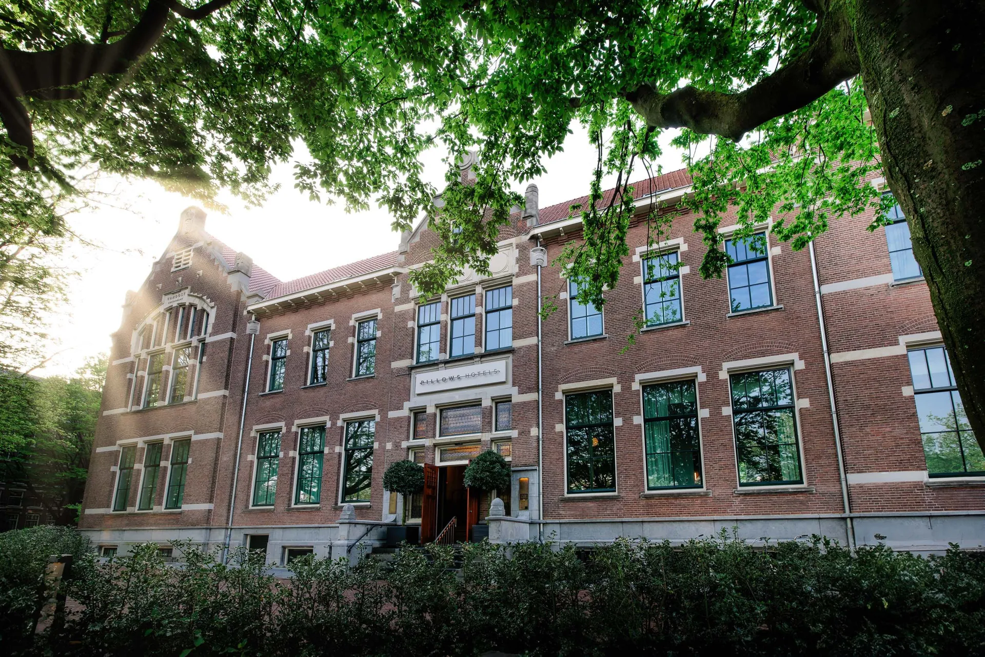 The red-brick facade of the hotel framed by tall park trees.