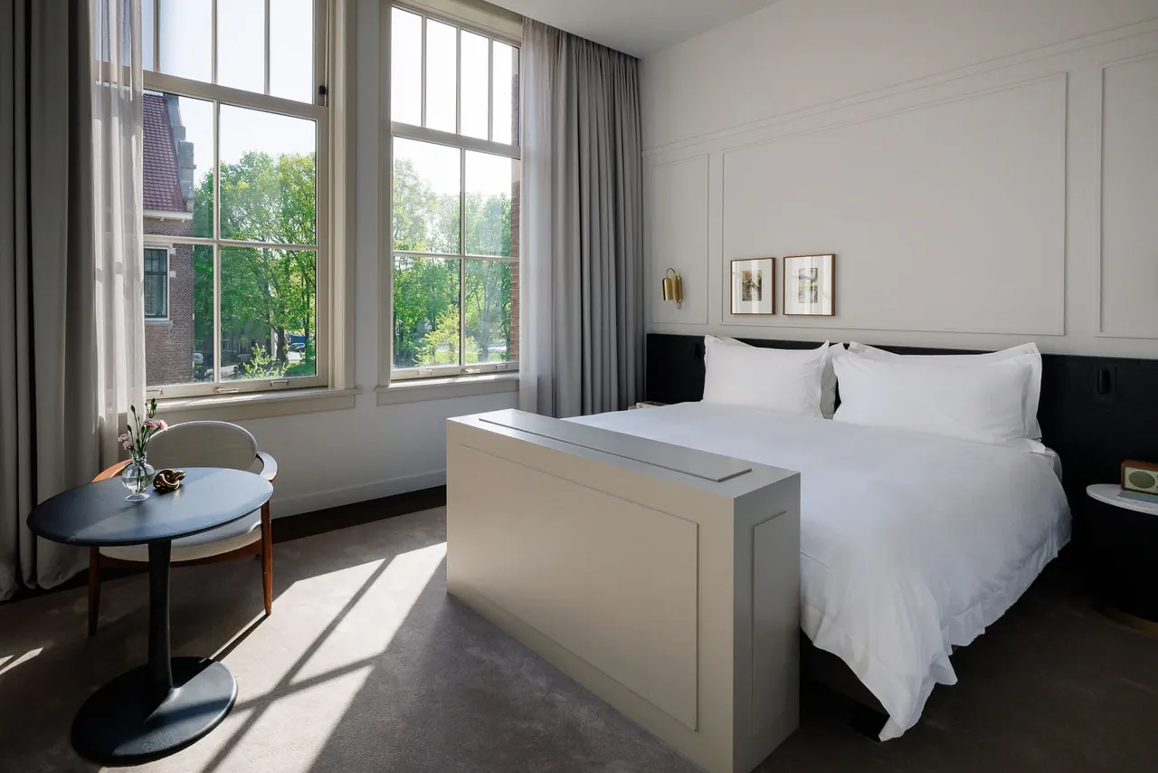 A suite with tall windows, a white bed and a small round table.