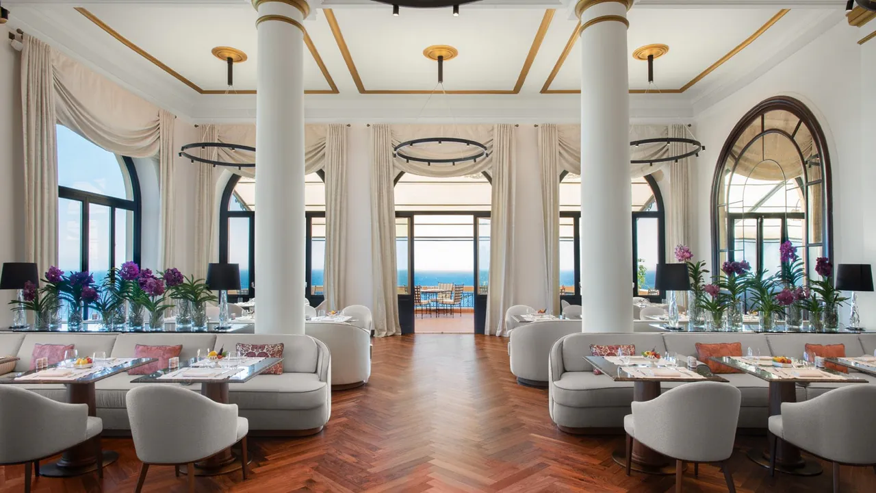 A bright dining room with white columns, arched windows and a glimpse of the sea.