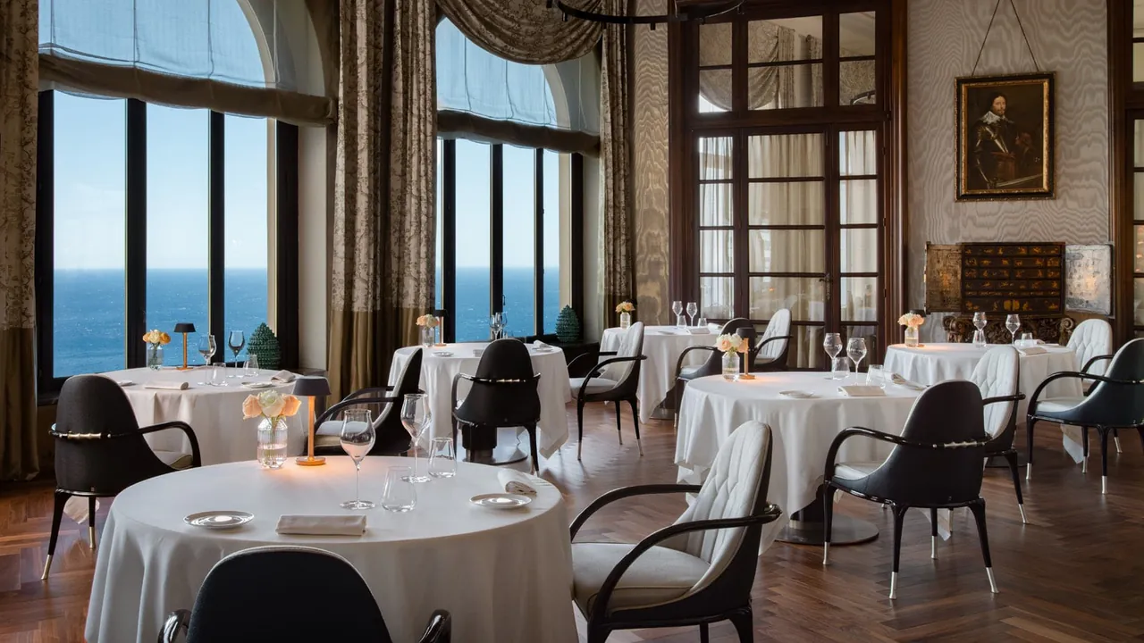 Round tables laid in a dining room with tall windows facing the sea.