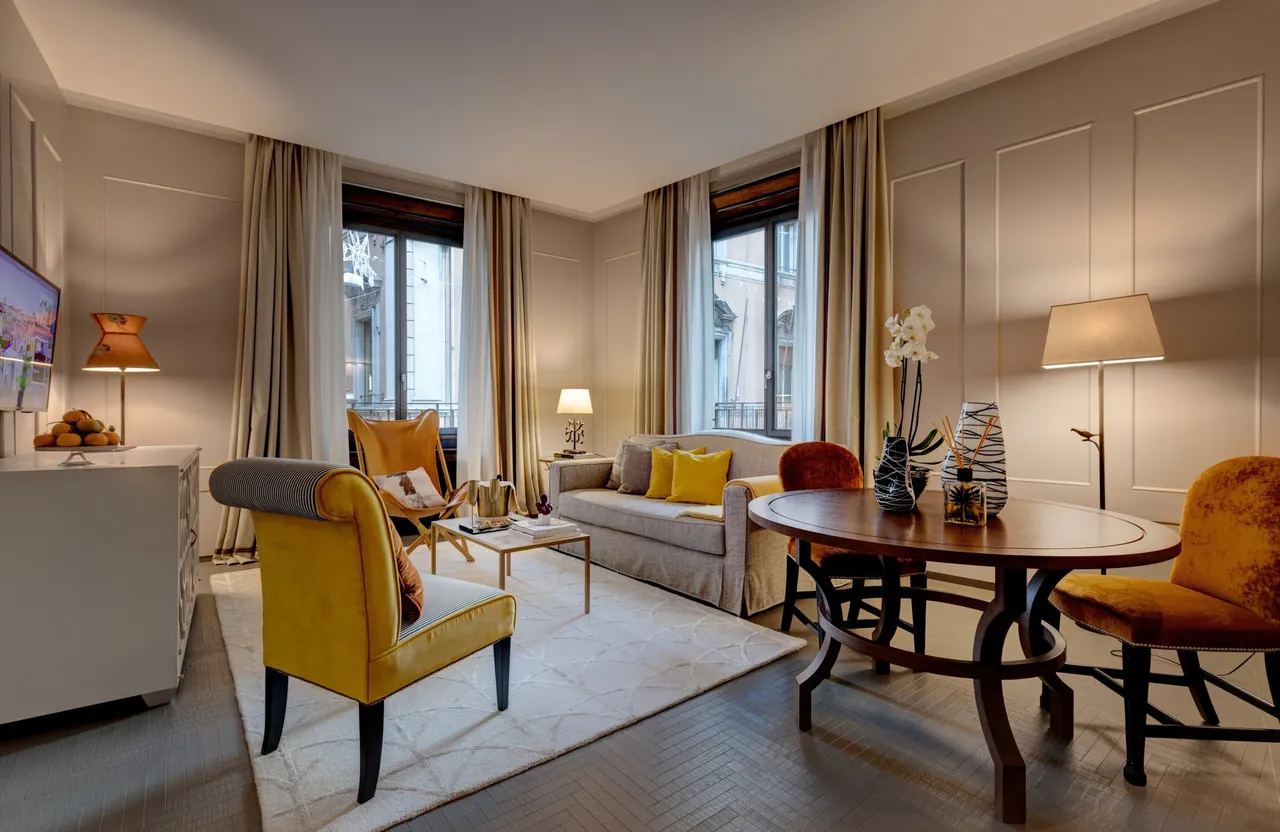 An apartment suite lounge with yellow armchairs, a round table and tall windows.