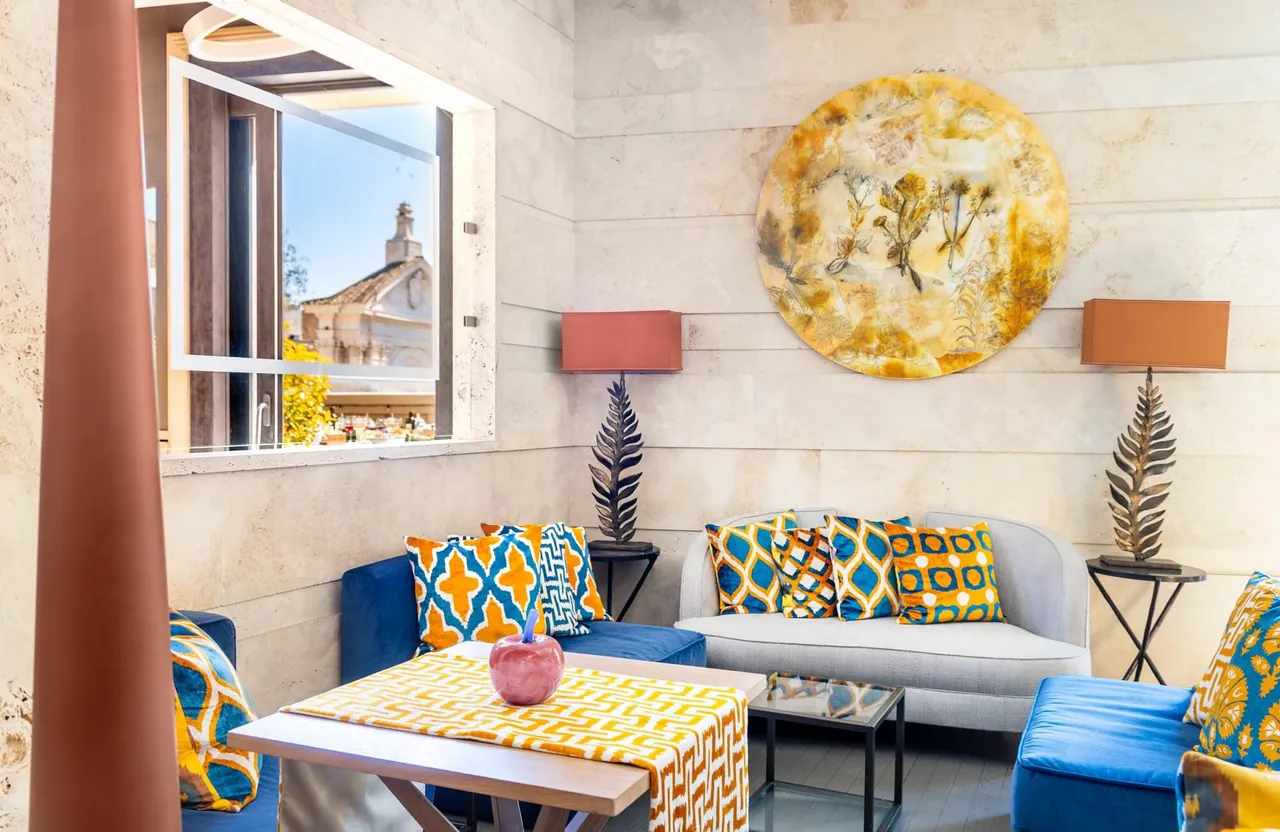 A bright living area with blue sofas, patterned cushions and a window over the rooftops.