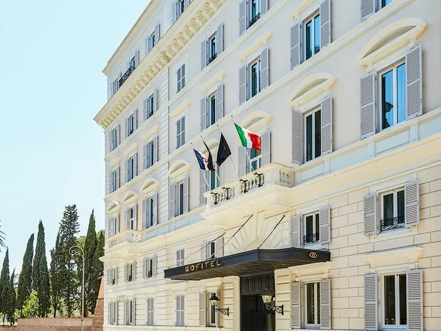 The cream facade of the hotel with flags above the entrance and cypress trees beside it.