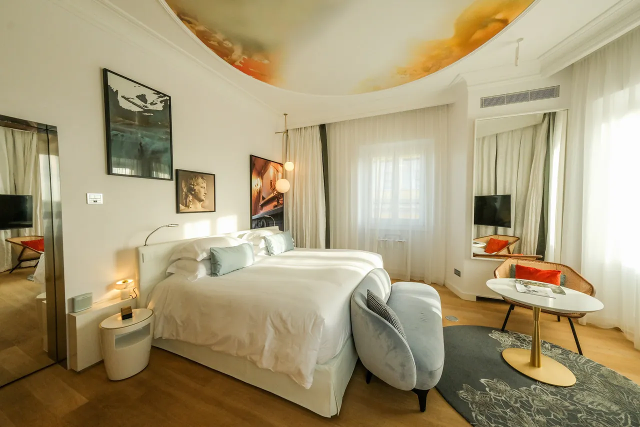 A bright guest room with a large white bed, a painted ceiling and a small seating corner.