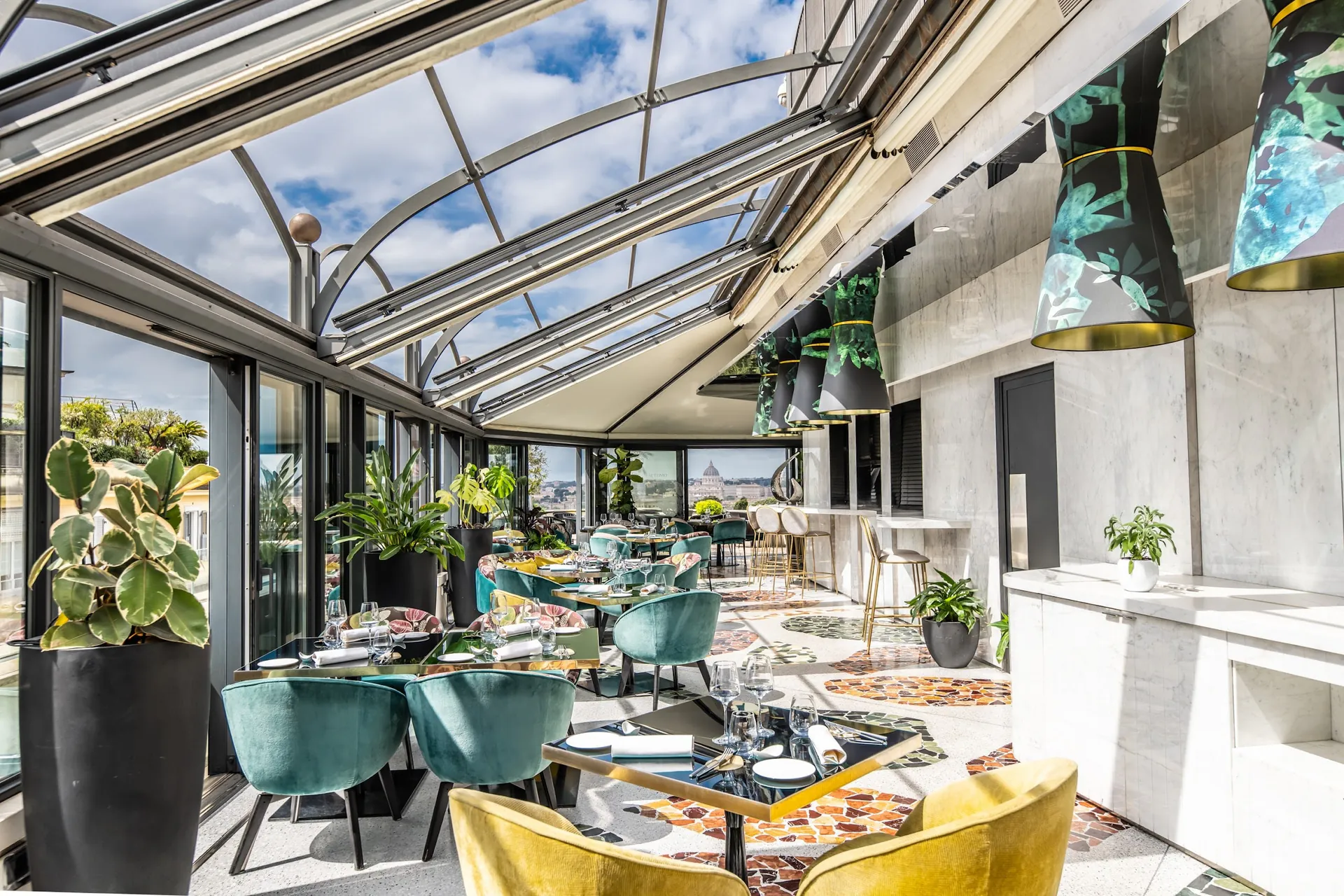 The glass-roofed rooftop restaurant with green chairs and plants along the windows.