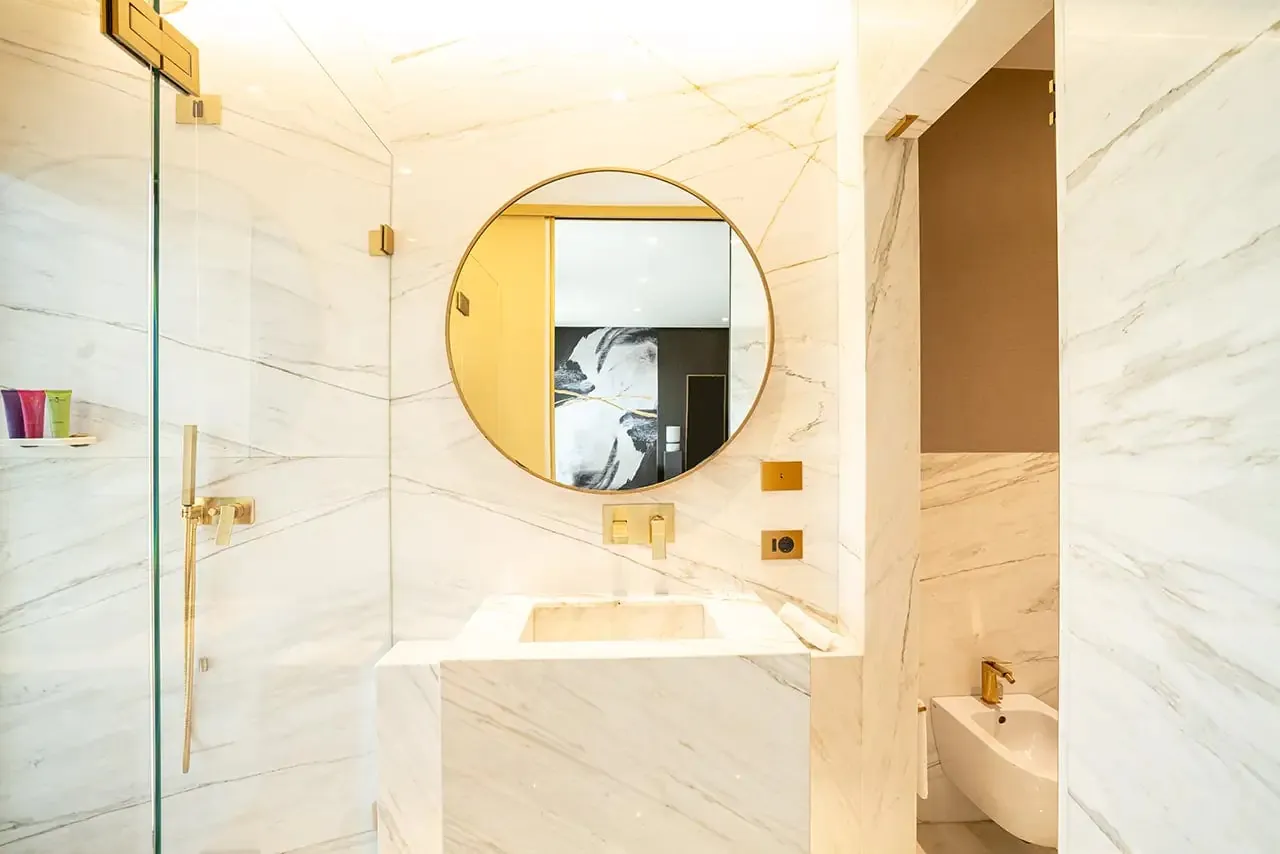 White marble bathroom with a round gold mirror and a glass shower.