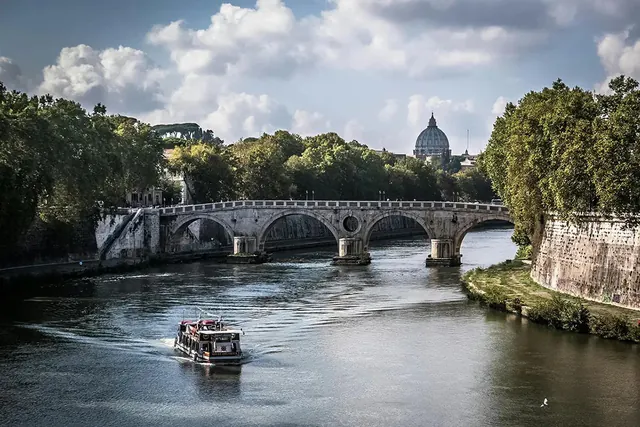 A tour boat on the Tiber with a stone bridge and St Peter's dome behind.