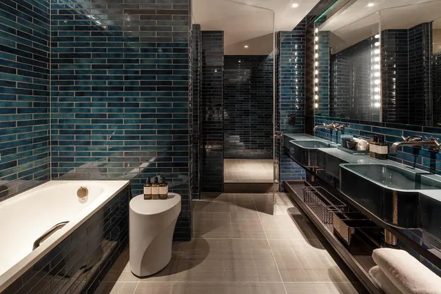 A bathroom lined with dark blue tiles, a bathtub and a lit vanity mirror.