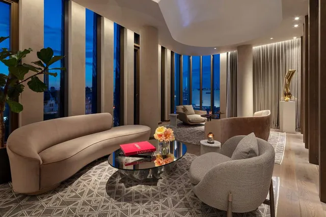 A suite living room with curved sofas and tall windows over the city at dusk.