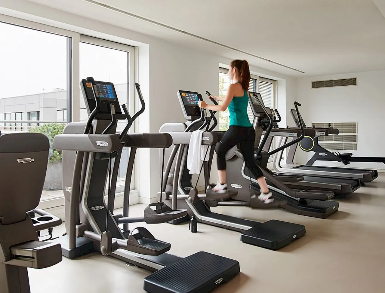 A fitness room with cardio machines and big windows.